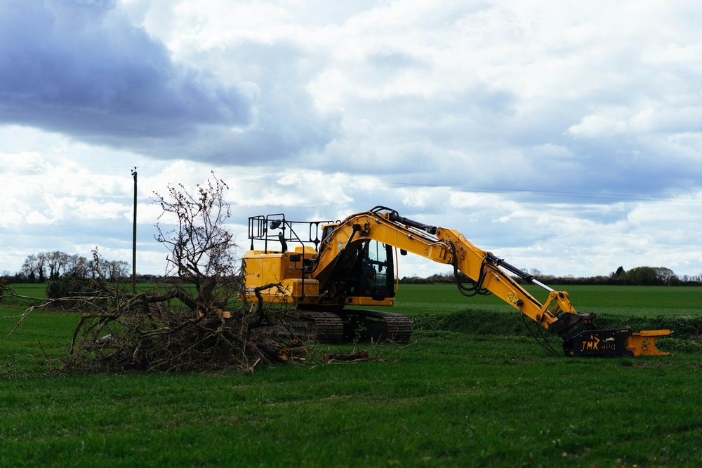An excavator with a grapple clearing cut branches at the edge of a field